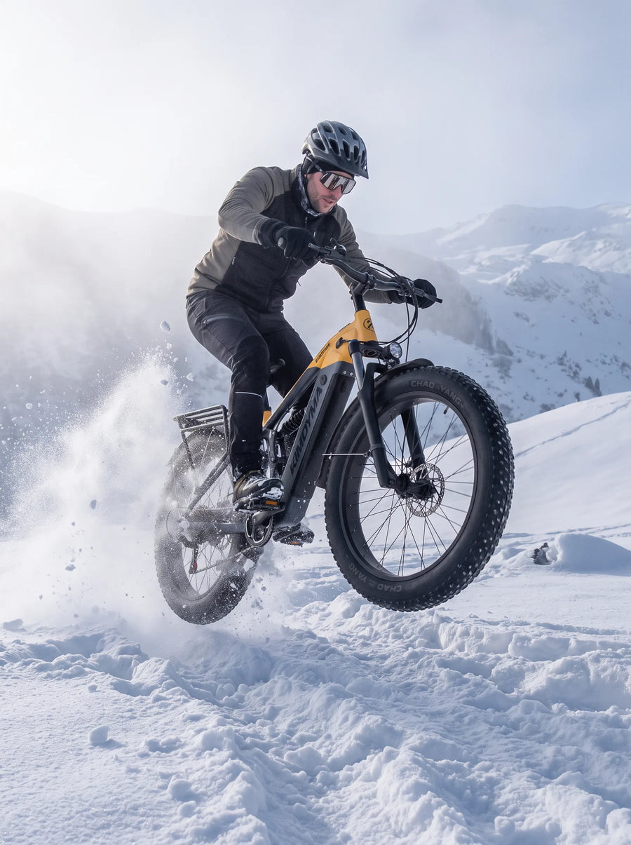Rowerzysta na rowerze elektrycznym Kimdyma K05 z oponami typu fat tire zeskakuje ze śnieżnego zbocza, wzbijając chmurę śniegu.