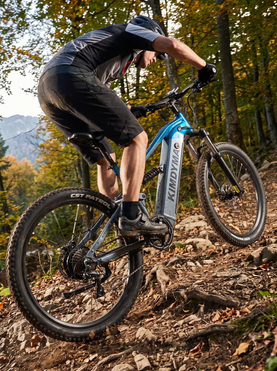 A cyclist riding a Kimdyma K01 electric mountain bike uphill on a dirt trail with gravel, surrounded by low shrubs, capt