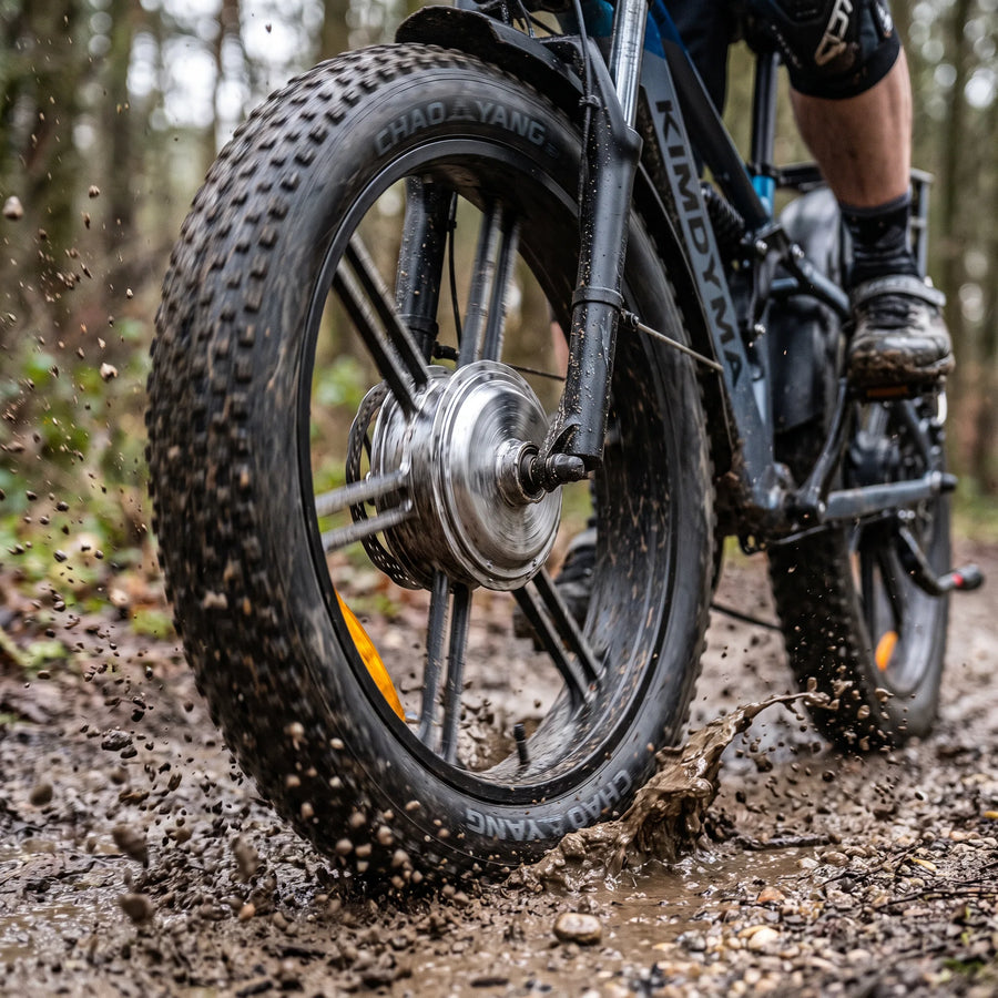 Close-up of Kimdyma K03's rear wheel with dual hub motor kicking up mud and gravel on a mountain trail, male rider's leg