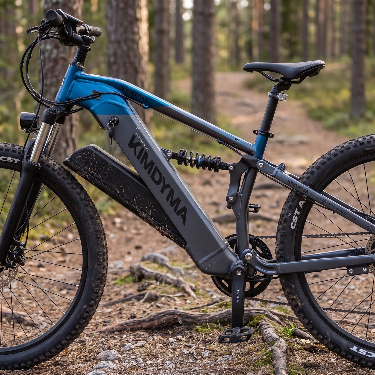 Una mountain bike elettrica Kimdyma blu e grigia con una grande batteria poggiata su un sentiero sterrato attraverso una pineta.